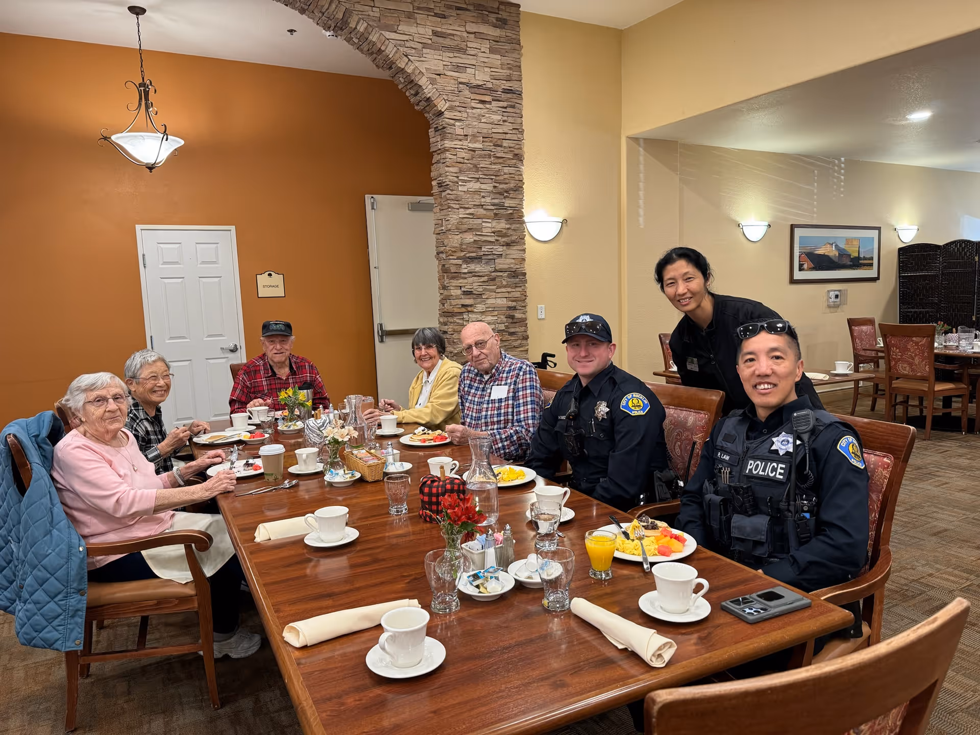 A group of elderly people and two police officers sitting around a wooden dining table in a senior living facility, enjoying a meal together. The room has warm lighting, a stone archway, and orange and beige walls. There are cups, plates with food, and glasses on the table.