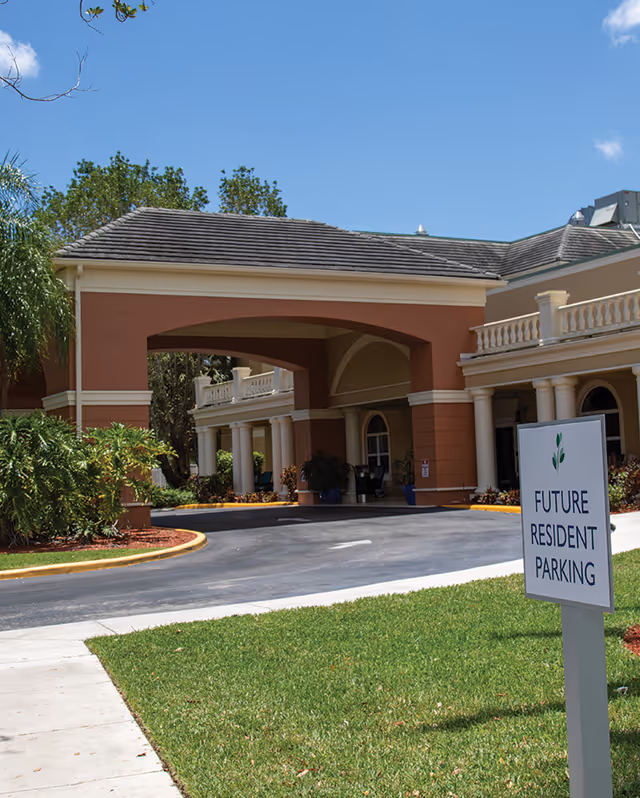 Entrance of a senior living facility with a covered driveway and columns, surrounded by greenery and a clear blue sky. A sign in the foreground reads 'Future Resident Parking'.