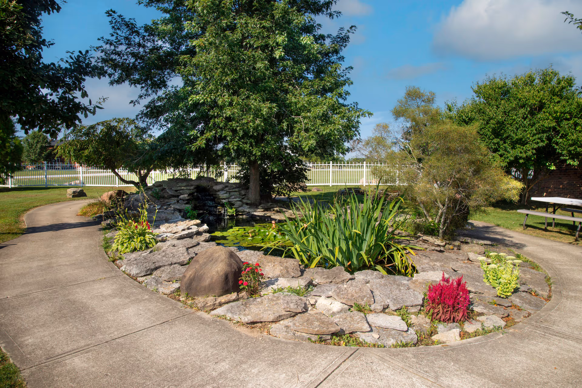 A landscaped outdoor garden area with a small pond surrounded by rocks, various plants, and trees. A curved concrete walkway encircles the garden, and there is a white fence and a picnic table visible in the background under a blue sky with some clouds.