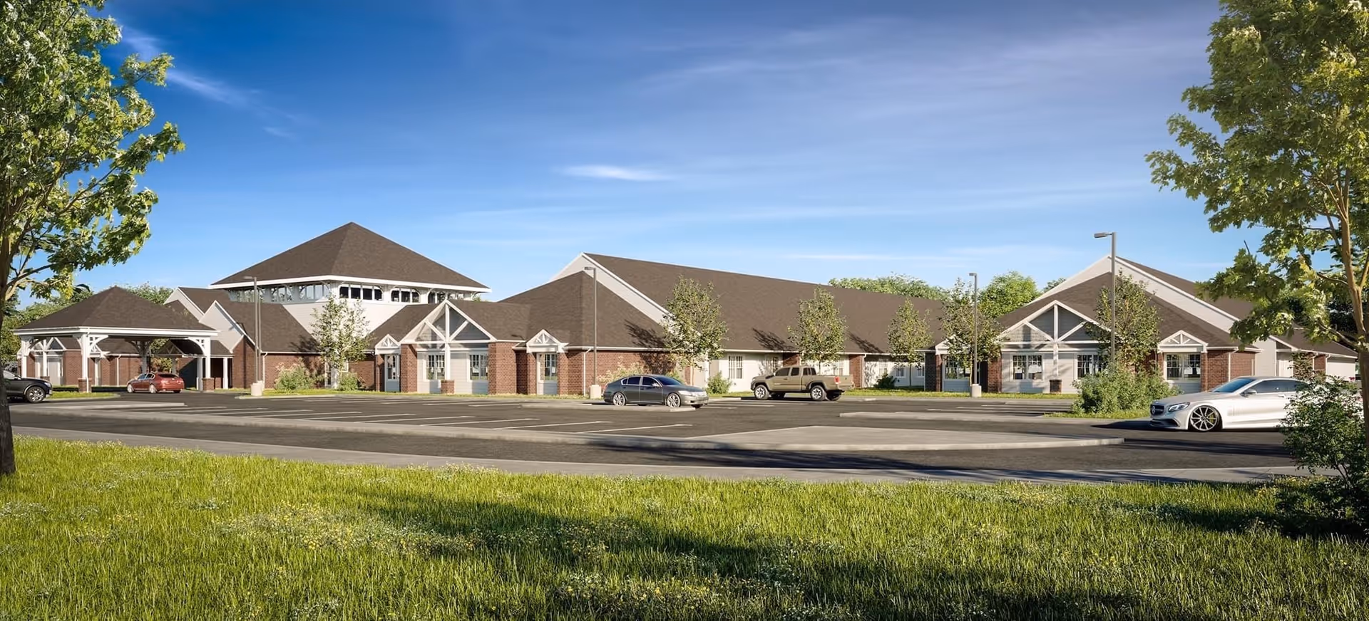 Low-rise brick senior living facility building with a parking lot, trees, and a grassy foreground under a blue sky.