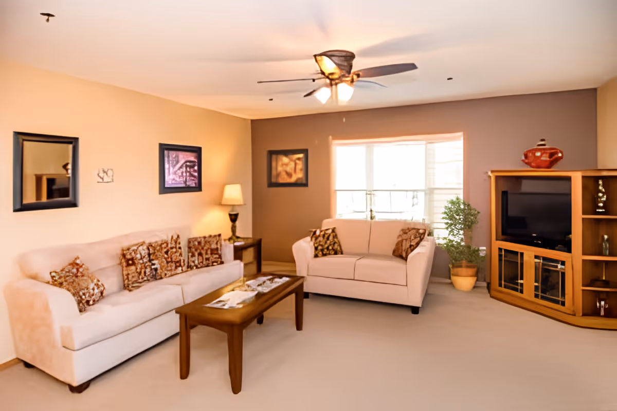 A cozy living room with two beige sofas adorned with patterned cushions, a wooden coffee table with magazines, a wooden entertainment center with a TV and decorative items, a potted plant near a window with blinds, a table lamp on a side table, and framed artwork on the walls. The ceiling has a fan with lights.