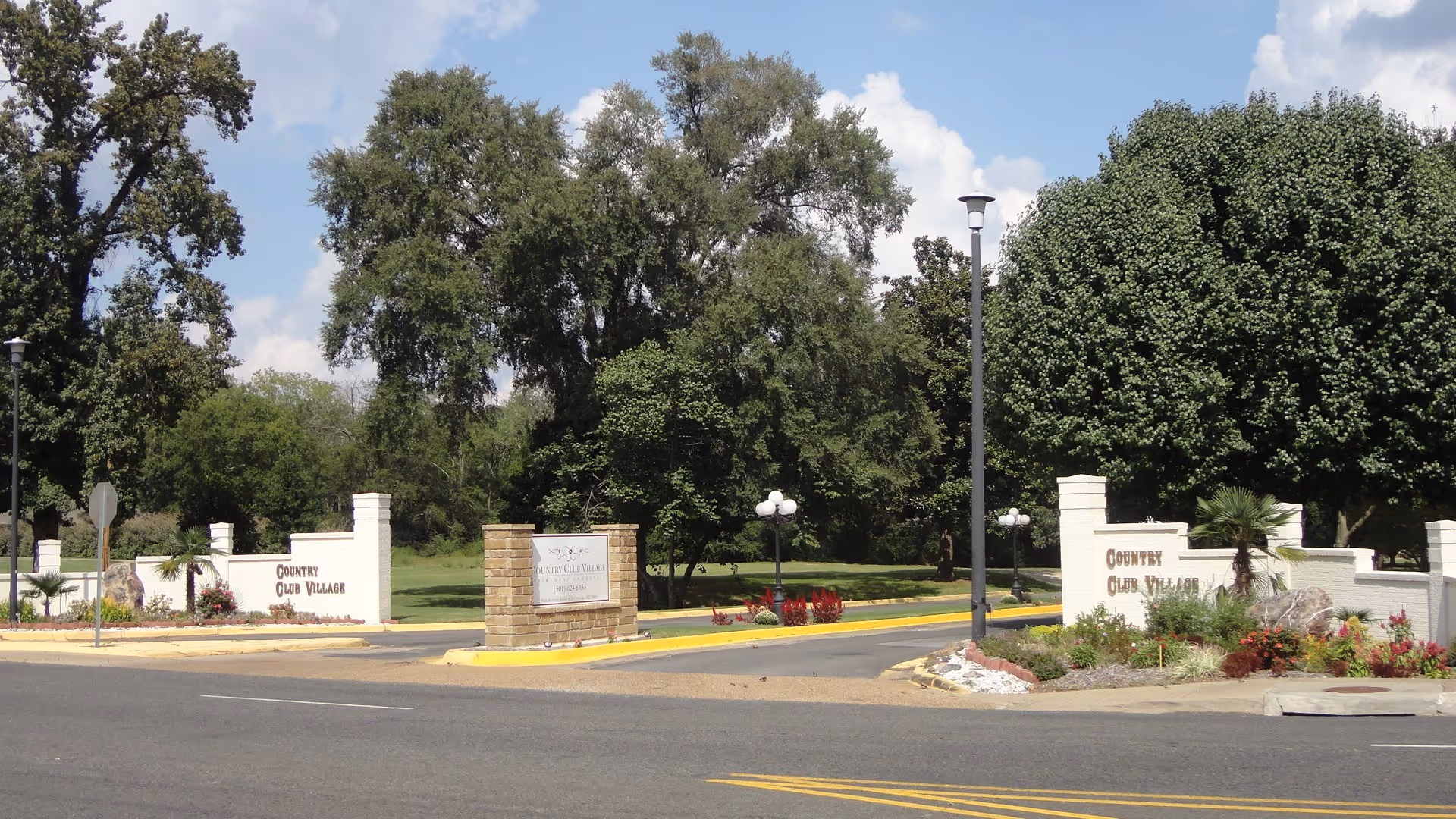 Entrance to Country Club Village Retirement Community featuring a wide driveway flanked by white brick walls with the community name on both sides, surrounded by landscaped greenery and large trees under a partly cloudy sky.