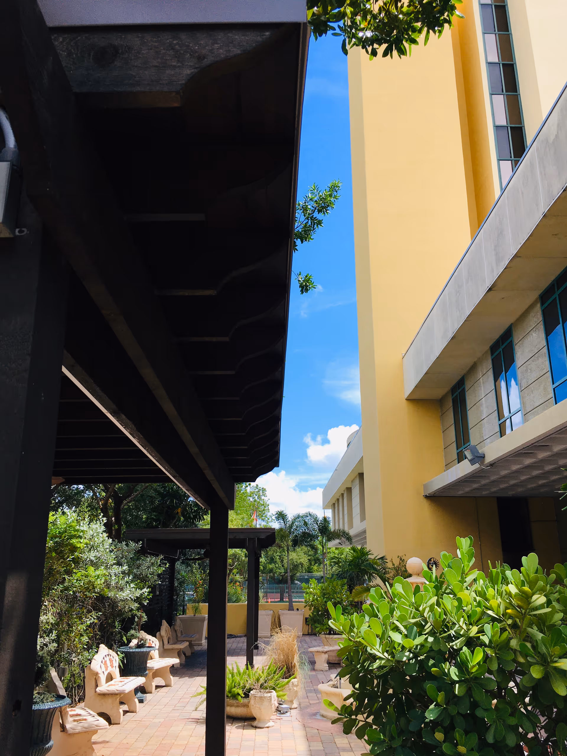 Outdoor patio area at Victoria Nursing & Rehabilitation Center with benches, potted plants, and a covered walkway. The building is yellow with large windows, and there are trees and greenery surrounding the space under a clear blue sky.
