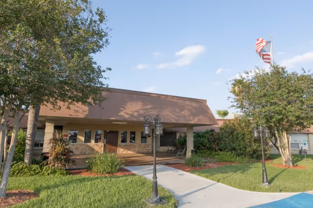 Exterior front entrance of a single-story rehabilitation facility with a walkway, lawn, lamp posts, and an American flag.