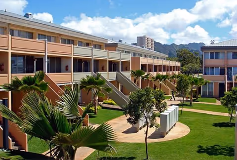 Exterior view of a tan senior living complex with balconies, palm trees, and a landscaped courtyard.
