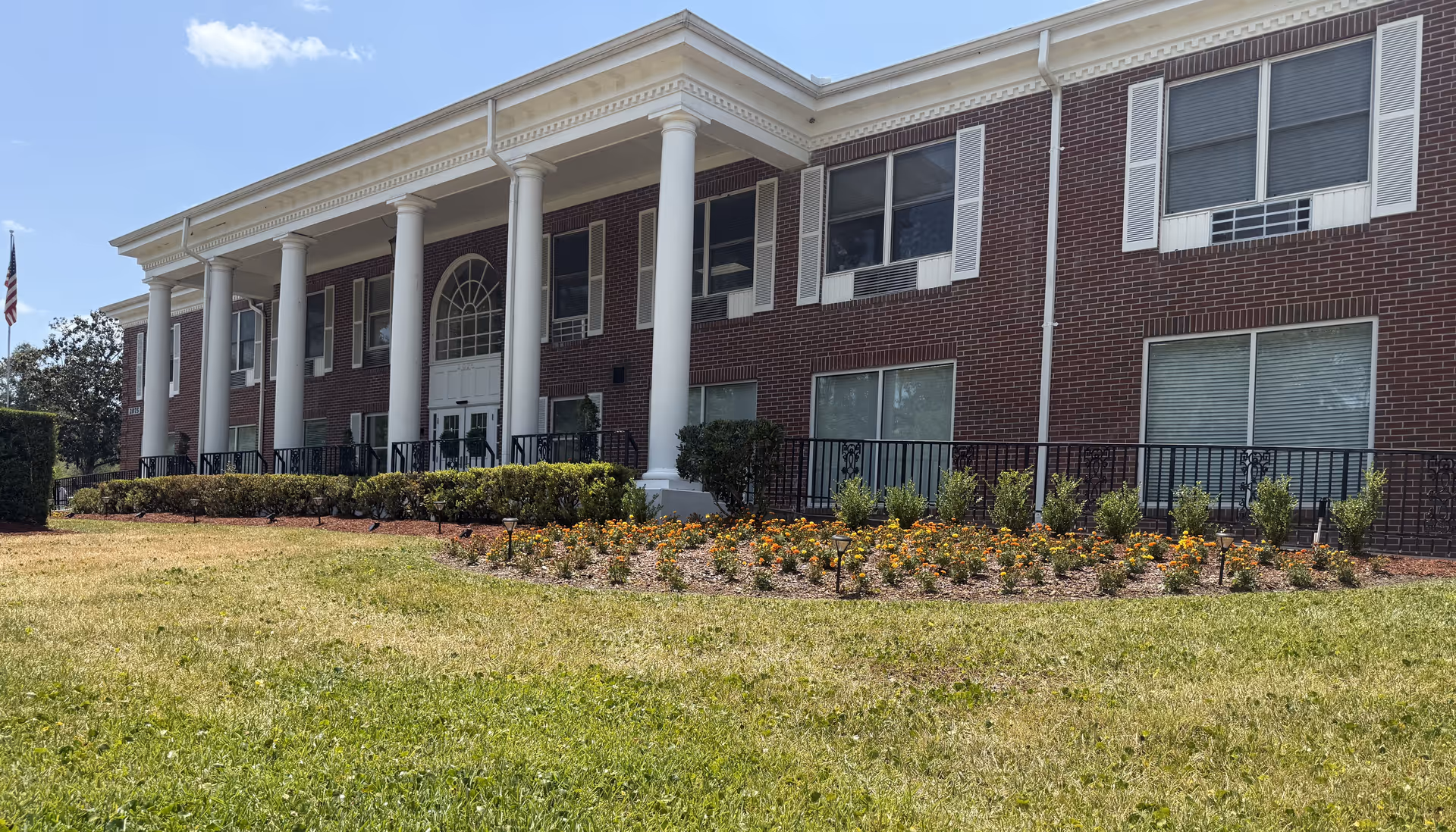 Exterior view of a two-story brick building with white columns and multiple windows. The building is surrounded by a well-maintained lawn and flower beds with yellow and orange flowers. An American flag is visible on the left side of the image under a clear blue sky.