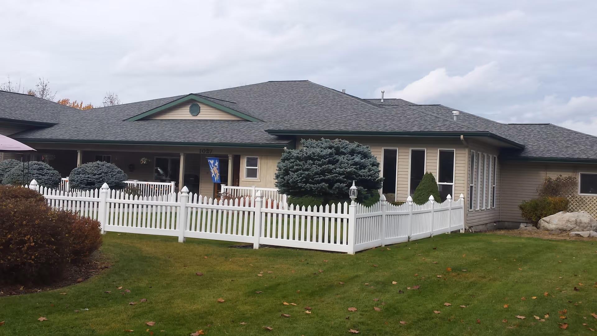 Front exterior of a single-story assisted living building with a white picket fence, lawn, and shrubs.