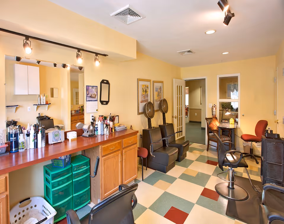 Interior view of a hair salon area in a senior living facility with a long counter holding various hair care products, two black salon chairs with hair dryers, a small desk with a lamp and chair, and a checkered floor with green, beige, and red tiles.