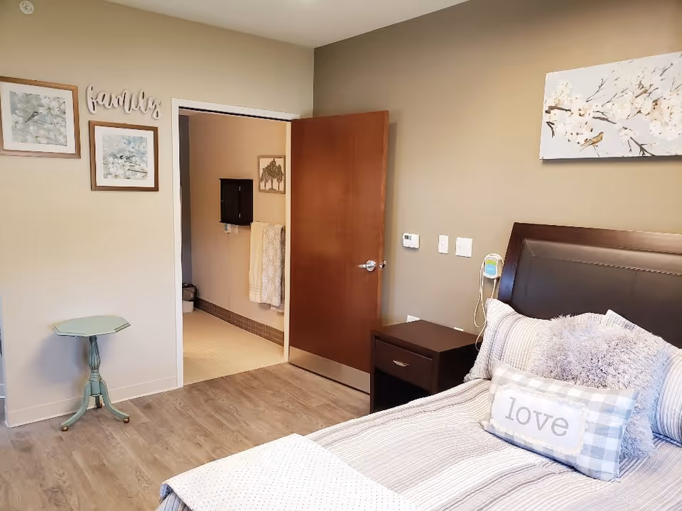 A cozy bedroom with a neatly made bed featuring pillows, including one with the word 'love'. The room has light-colored walls decorated with framed floral artwork and a wooden door leading to a bathroom. There is a small green side table and a dark wooden nightstand beside the bed.