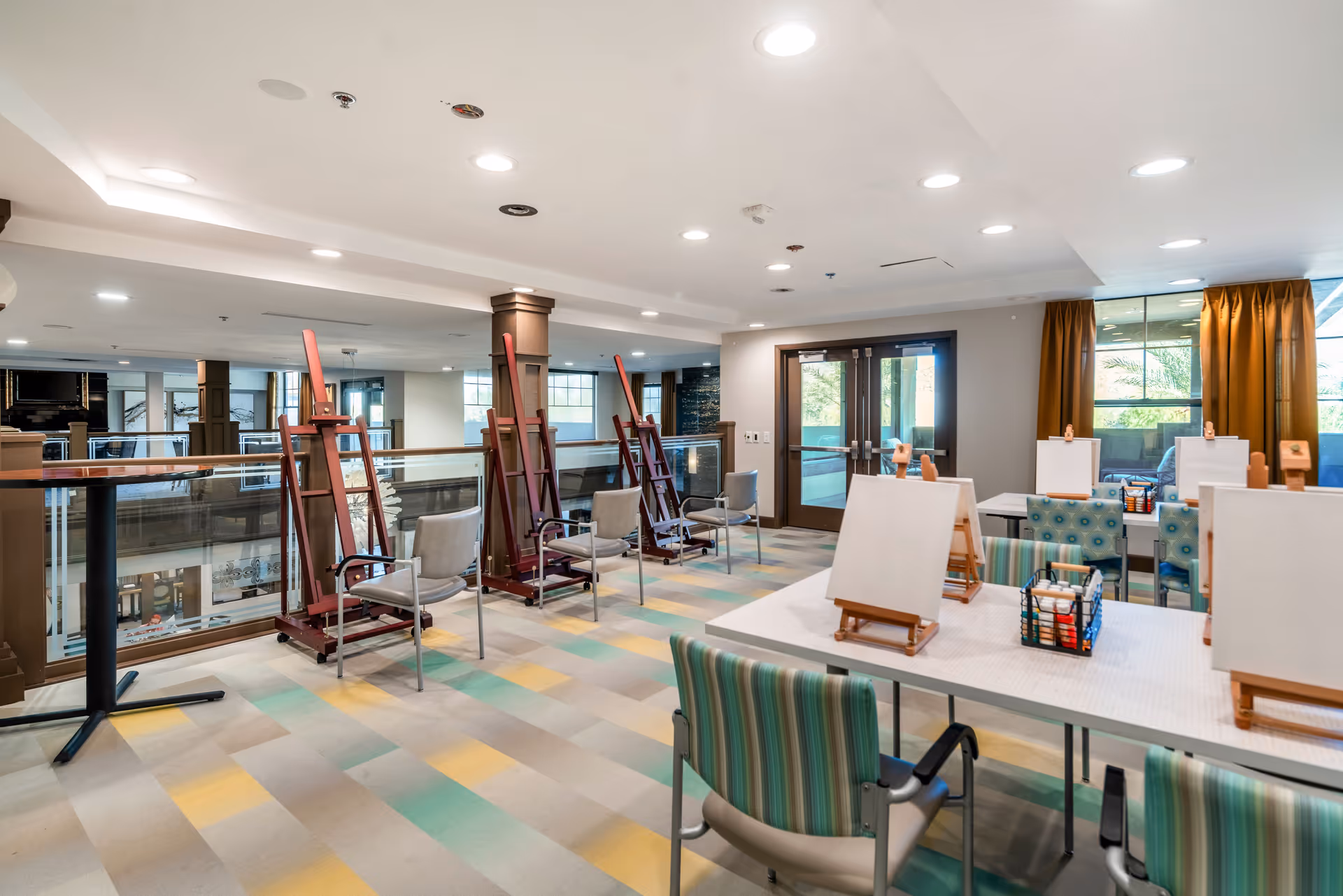 A bright and spacious art studio area in a senior living facility with multiple easels and chairs arranged around tables. The room has large windows with yellow curtains allowing natural light to enter, and the floor is covered with a colorful patterned carpet. The space is well-lit with ceiling lights and has a modern, clean design.