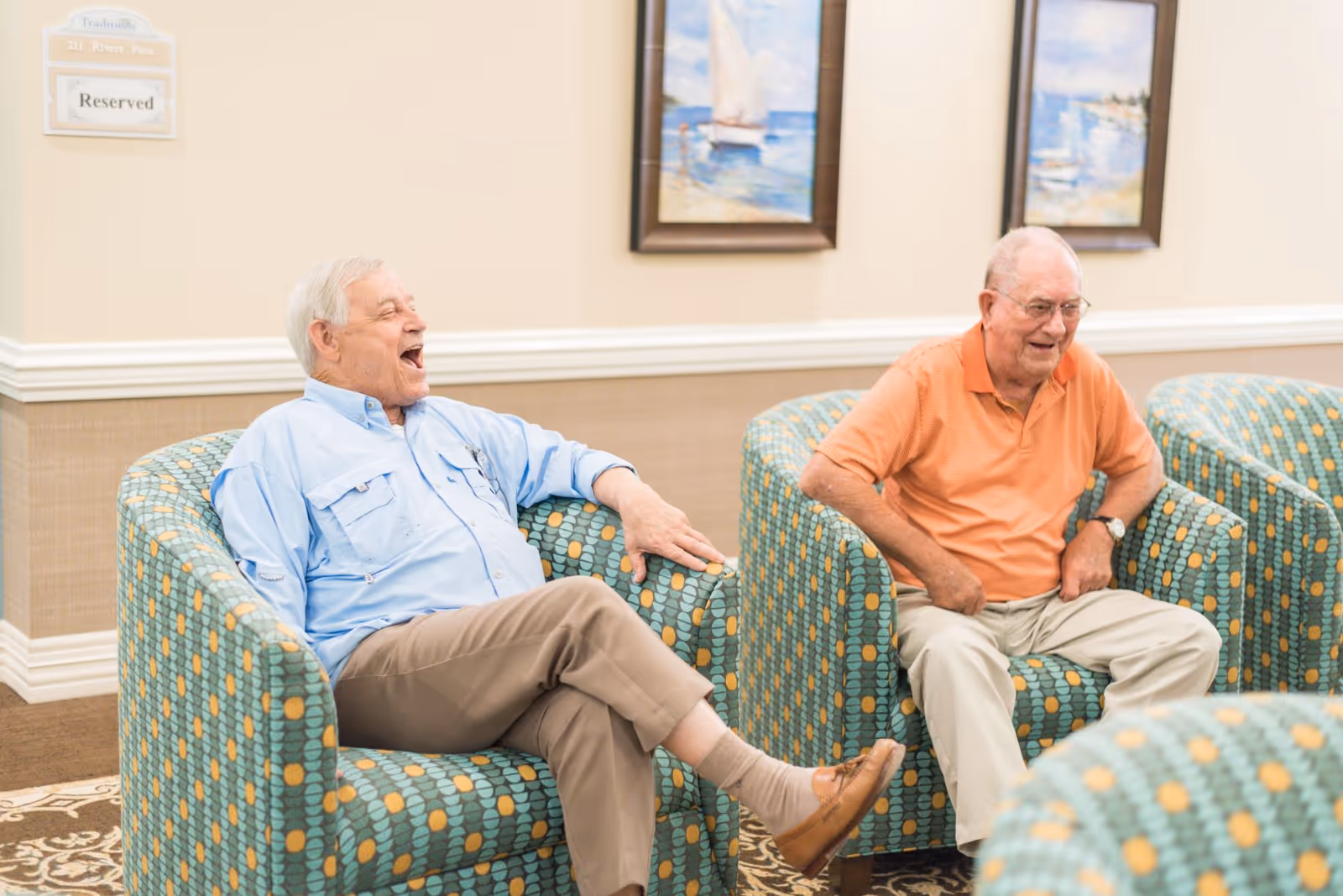 Two elderly men sit and laugh in patterned armchairs in a senior living facility lounge.