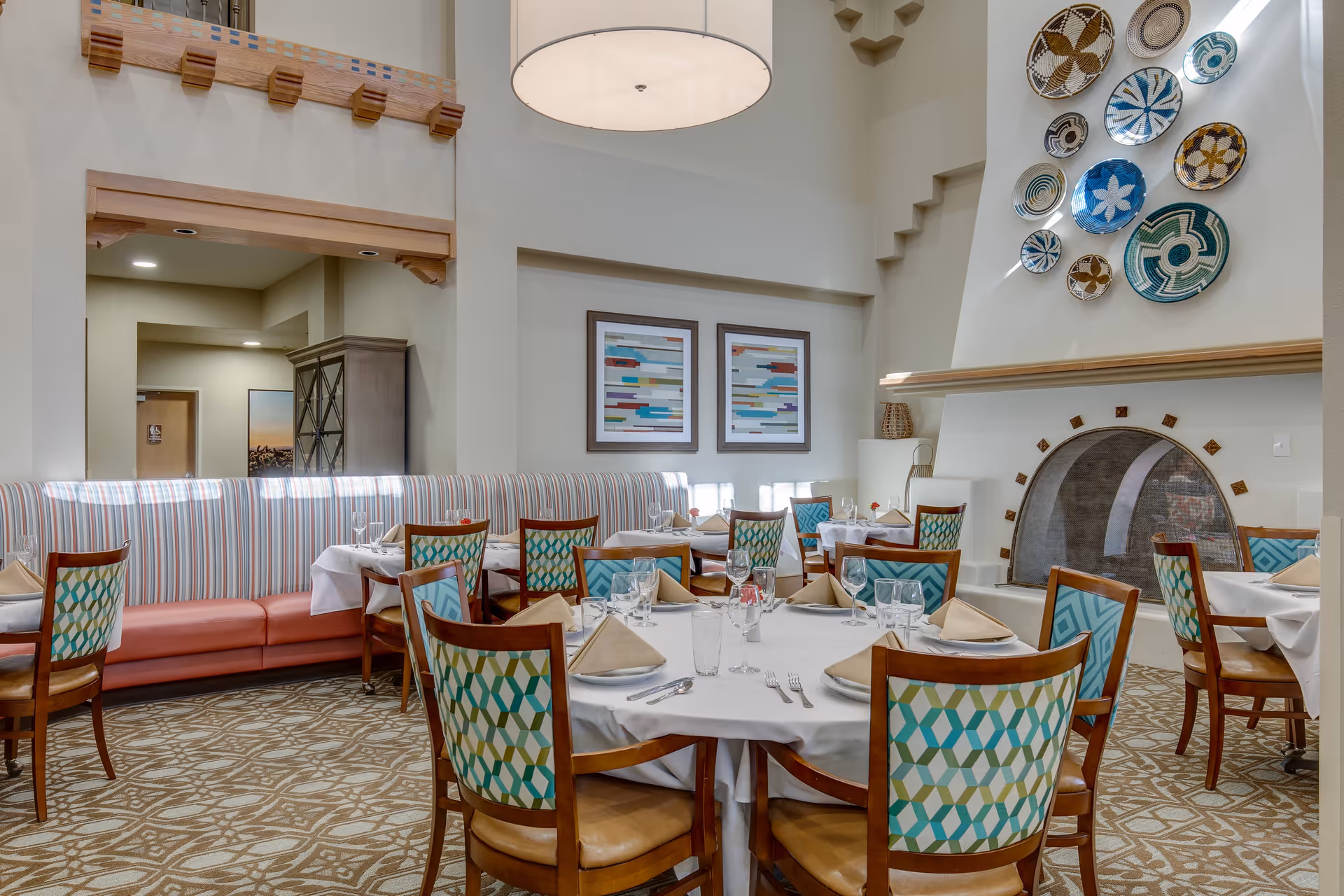 A dining room with round and rectangular tables set with white tablecloths, beige napkins, glassware, and silverware. The chairs have wooden frames with patterned upholstery in shades of blue, green, and beige. There is a large fireplace with decorative plates mounted above it on the wall. The room has high ceilings and neutral-colored walls with framed abstract artwork.