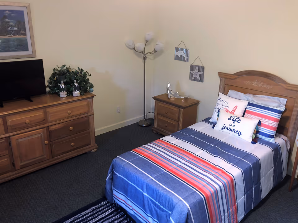 A bedroom with a single bed covered in a striped blue, red, and white bedspread. The bed has decorative pillows with phrases like 'Life is a journey' and 'Relax you're on sea time.' Next to the bed is a wooden nightstand with a silver dolphin figurine. A floor lamp with five white shades stands in the corner. Across from the bed is a wooden dresser with a flat-screen TV, two small decorative bottles, and a green plant. A framed picture hangs on the wall above the dresser.
