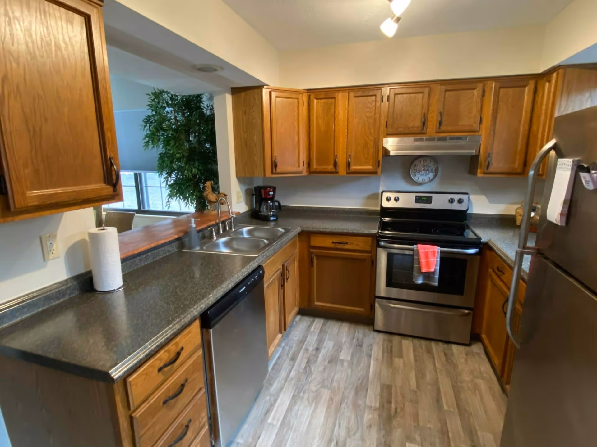 A kitchen with wooden cabinets, a stainless steel dishwasher, stove, and refrigerator. The countertop is dark and there is a double sink with a faucet. A coffee maker and a paper towel roll are on the counter. The floor has a wood-like finish and there is a window with a view of a green plant and a dining area beyond.