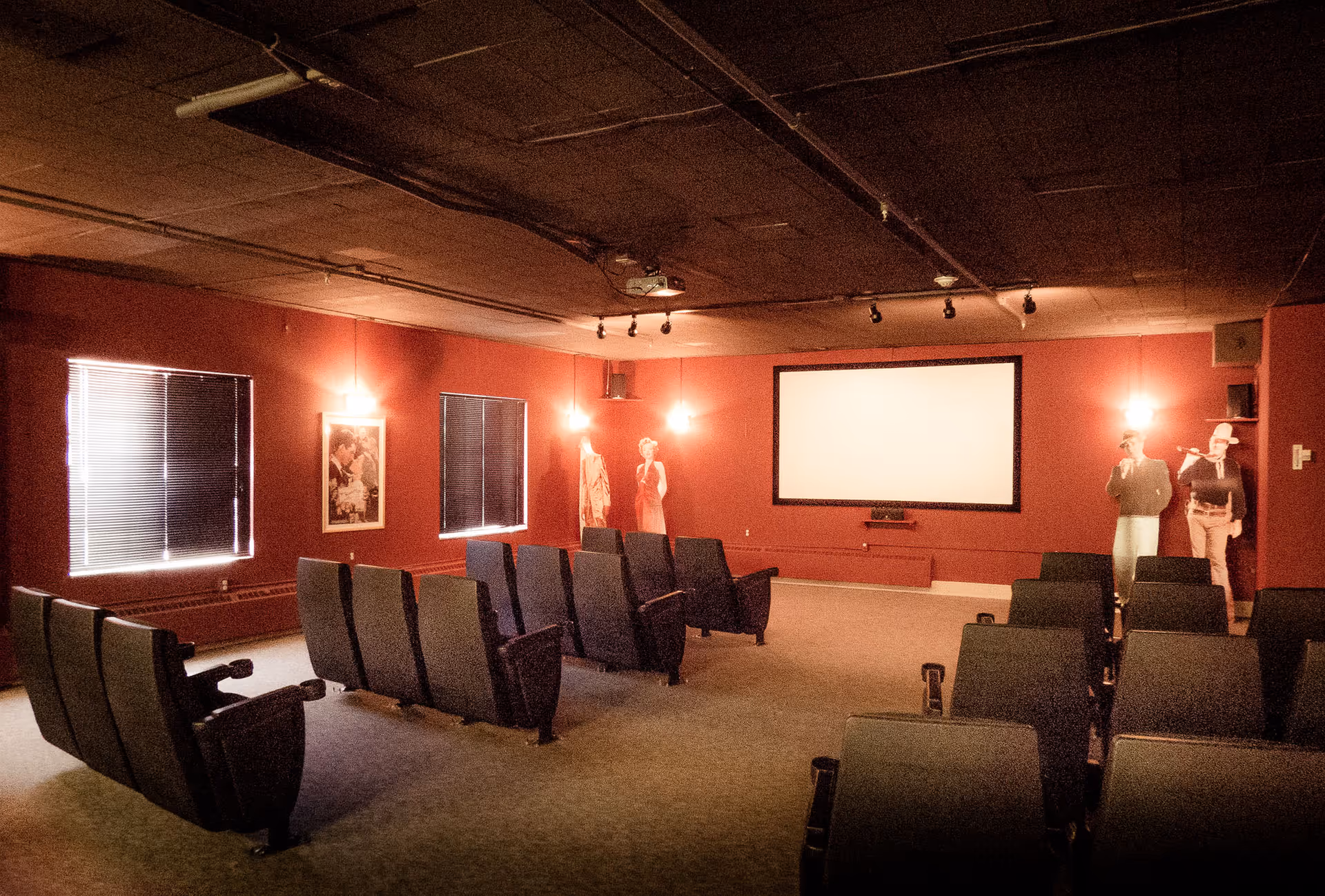 Small theater-style screening room with rows of seats facing a projection screen on red walls under dim lighting.