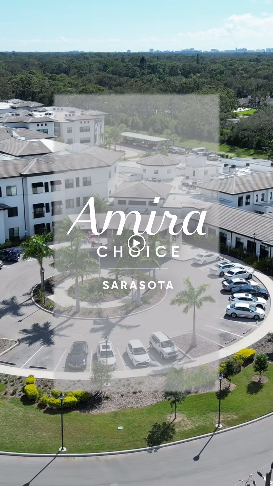 Aerial view of the Amira Choice Sarasota senior living facility showing multiple white buildings with gray roofs, a circular driveway with parked cars, palm trees, and surrounding greenery.