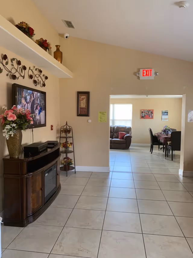Tile-floored interior hallway with a TV cabinet and floral decorations leading into a seating area and dining table under an exit sign.