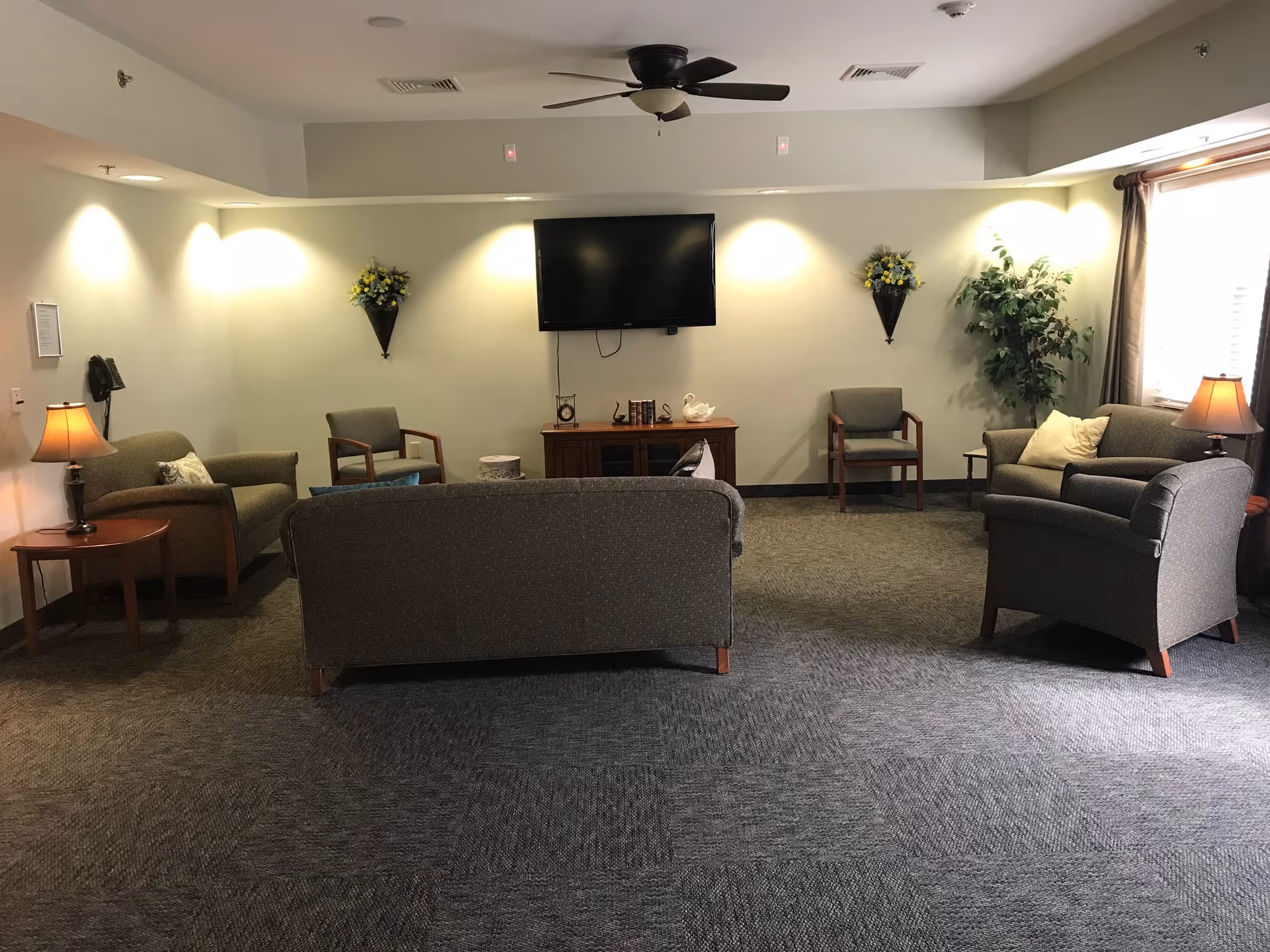 A cozy living room area with gray upholstered sofas and chairs arranged around a wall-mounted flat screen TV. The room has carpeted flooring, two wall-mounted flower arrangements, a potted plant near a window with curtains, and two table lamps on side tables providing warm lighting.