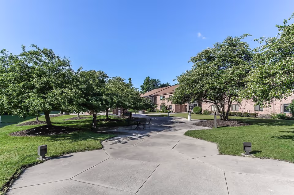 A paved walkway surrounded by green grass and trees leading to a two-story brick building under a clear blue sky. There are benches and a table with chairs along the path.