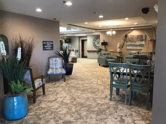 Carpeted communal lounge in a retirement center with armchairs, a small dining table, potted plants, and wall art.