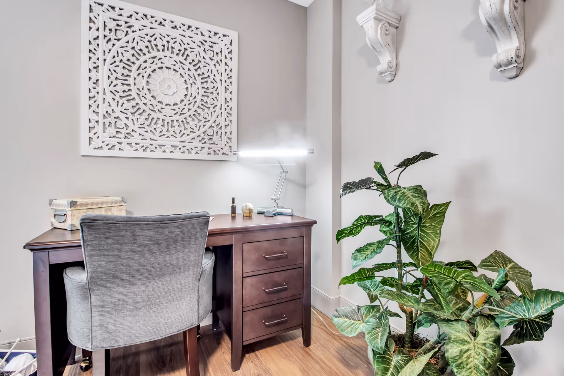 A small home office setup with a dark wooden desk and a gray upholstered chair. On the desk are a modern desk lamp, a decorative box, and small decorative items. Behind the desk is an intricate white wall art piece, and to the right is a large green leafy plant. The walls are light gray, and the floor is wooden.