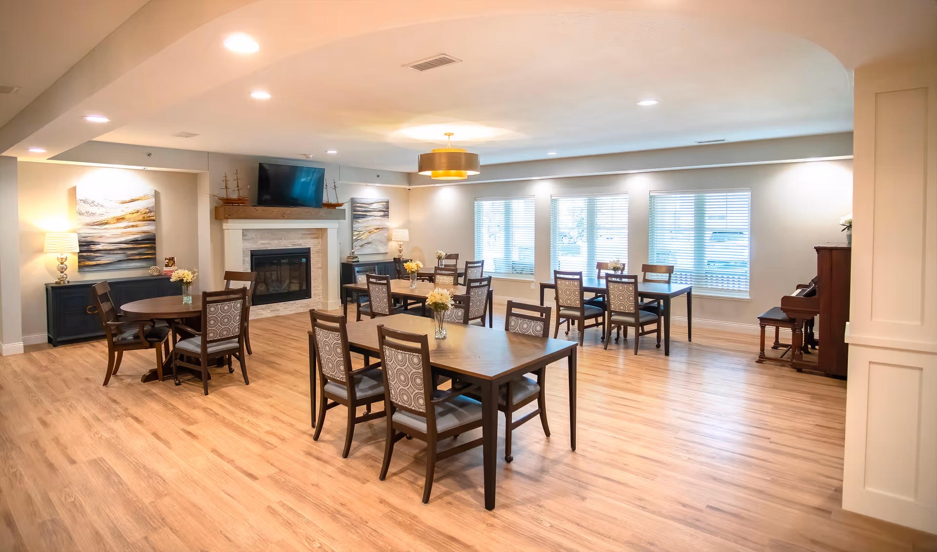 Bright communal dining/activity room with multiple tables and chairs, a fireplace with TV, large windows, and a piano.