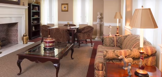 Cozy living room with a patterned sofa, armchairs, glass-top coffee table and fireplace, lit by table lamps.