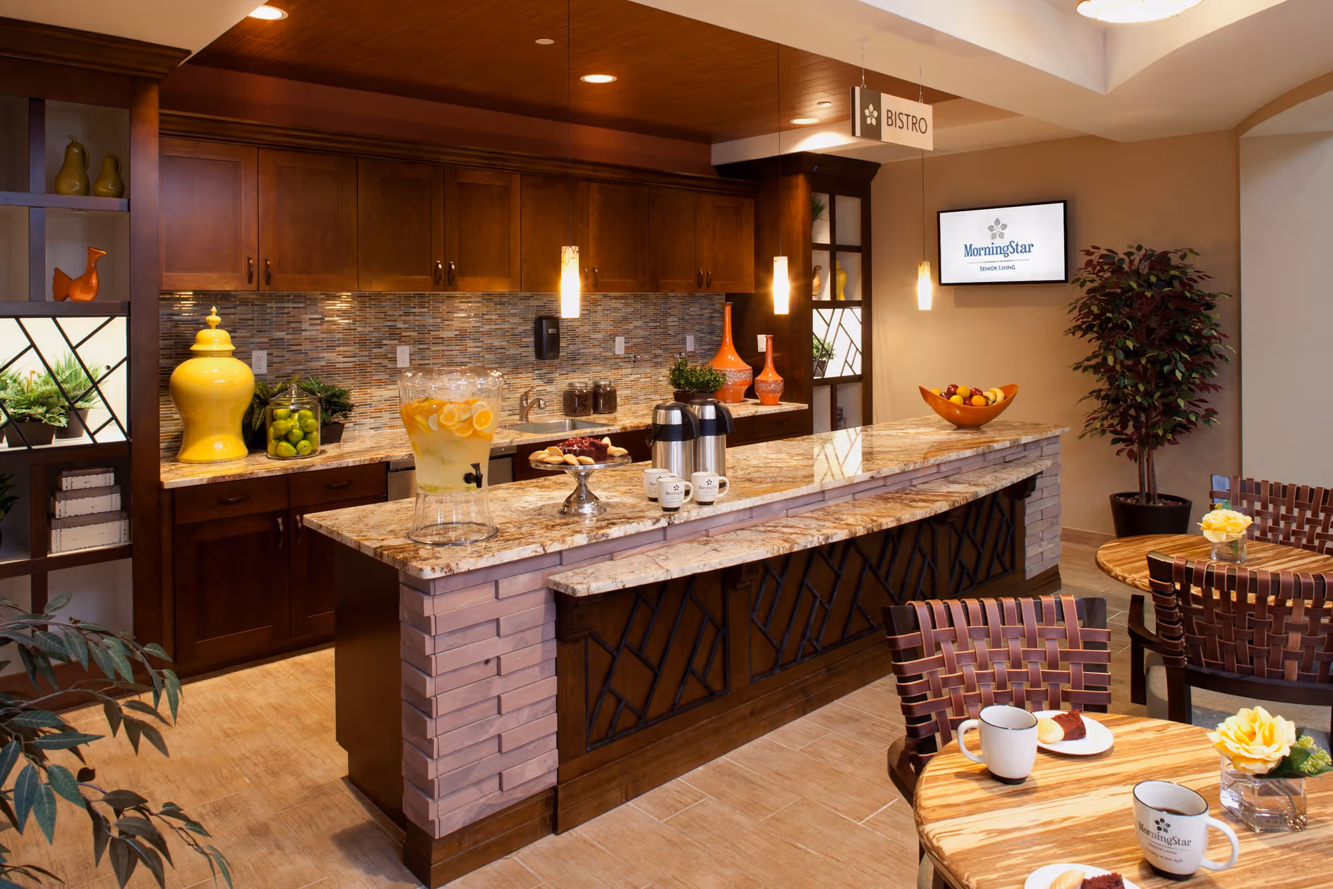 A cozy bistro area in MorningStar Assisted Living & Memory Care of Boulder featuring a granite countertop island with coffee pots, cups, and a glass dispenser filled with lemon water. The background shows wooden cabinets, decorative vases, and a mosaic tile backsplash. There are round wooden tables with woven chairs, each table adorned with a small flower arrangement and coffee cups. A wall-mounted screen displays the MorningStar Senior Living logo, and a sign hanging from the ceiling reads 'Bistro'.