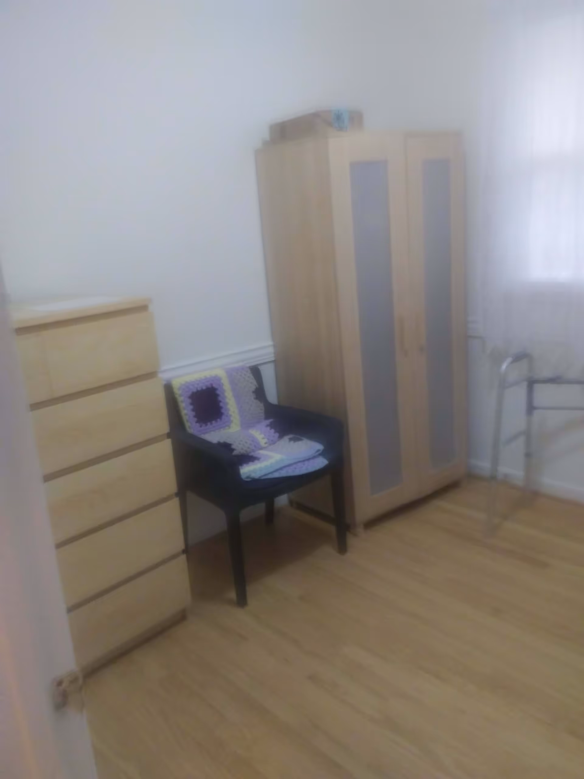 A small room with light wooden flooring and white walls. There is a light wooden chest of drawers on the left, a black chair with a crocheted blanket draped over it in the middle, and a light wooden wardrobe with frosted glass panels on the right. A metal walker is positioned near a window with sheer white curtains.
