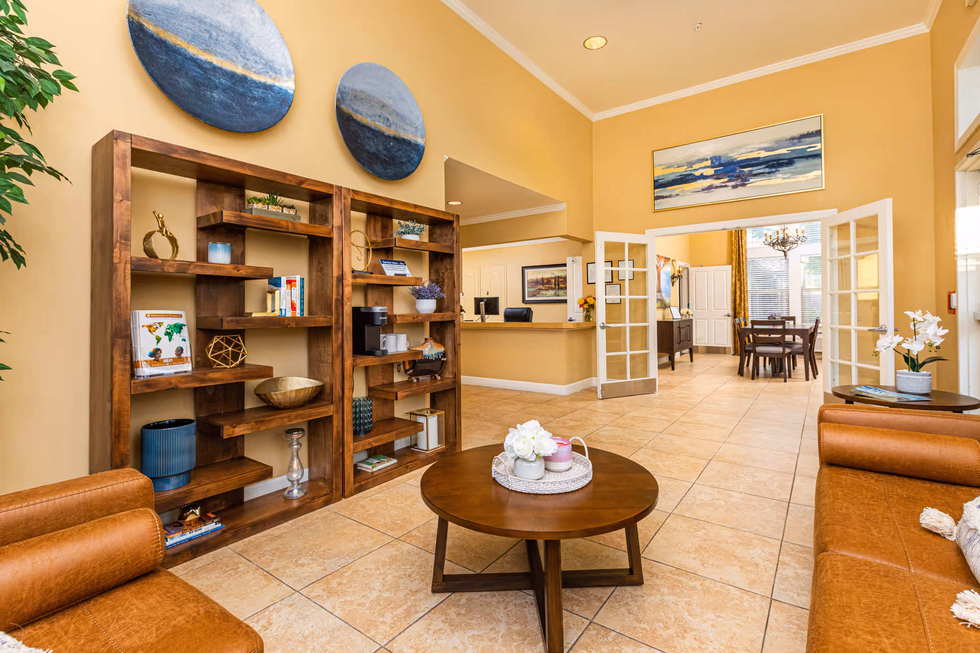A cozy senior living facility common area with tan walls and tiled floors. The room features two brown leather sofas facing a round wooden coffee table with decorative items on top. Behind the sofas is a wooden shelving unit with books, plants, and decorative objects. In the background, there is a reception desk and a dining area with a wooden table and chairs, illuminated by a chandelier. The space is warmly lit and decorated with abstract wall art and plants.