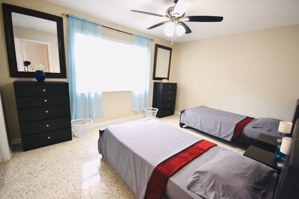 A bright bedroom with two single beds covered in gray bedding with red runners. The room has a ceiling fan with lights, two black dressers with mirrors above them, light blue curtains on the window, and two white laundry baskets on the floor.