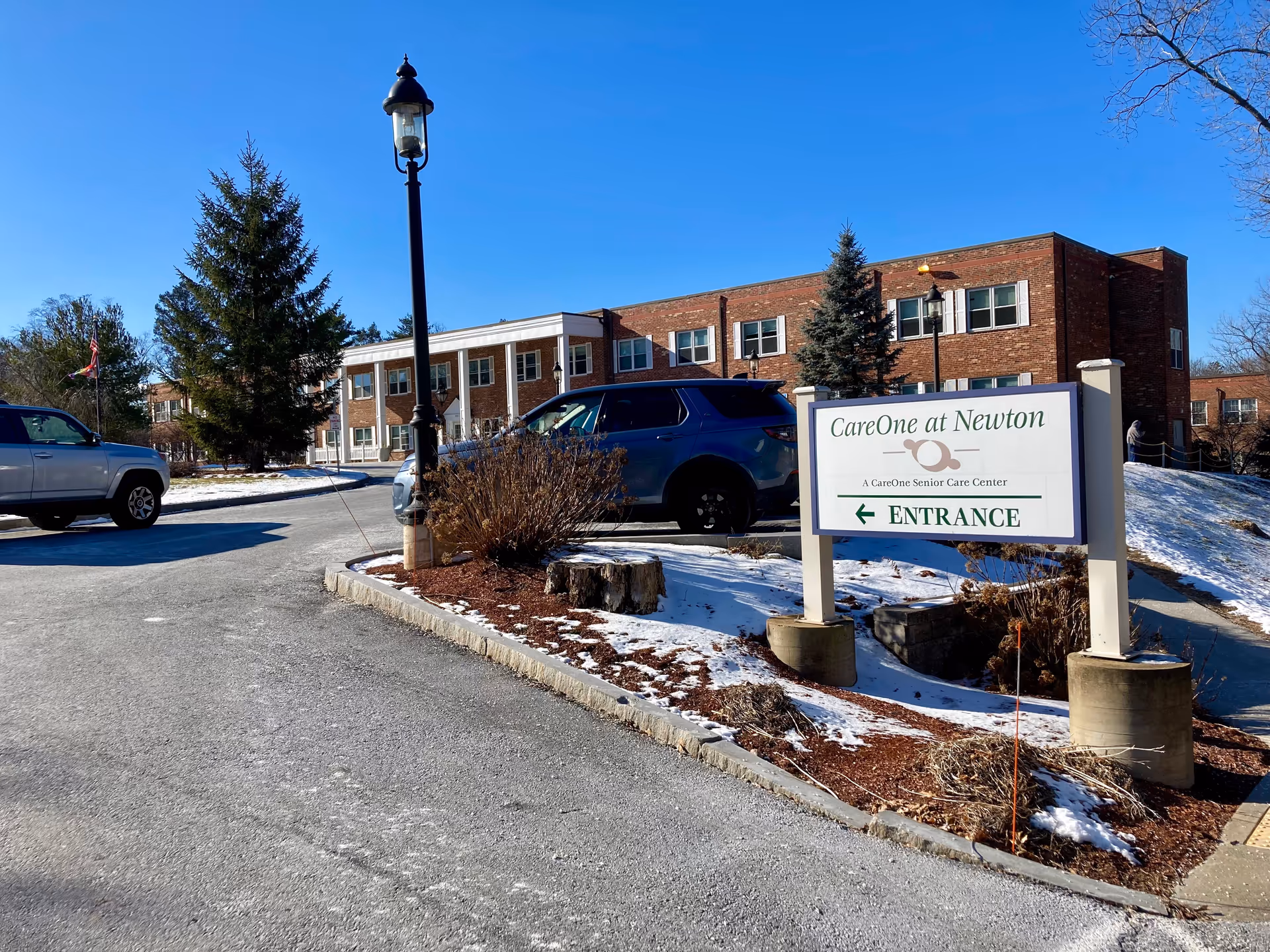 Exterior view of CareOne at Newton senior care center on a clear day with a blue sky. The image shows a brick building, a parking area with cars, a lamppost, some trees, and a sign indicating the entrance to the facility.
