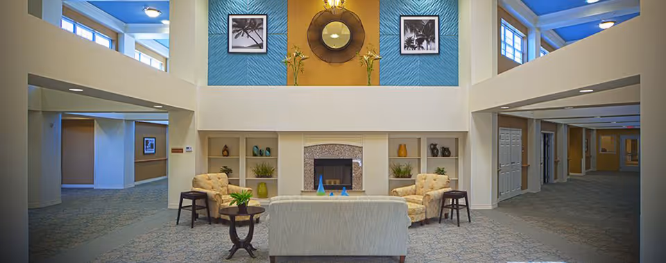 A spacious and well-lit common area in an assisted living facility featuring a central fireplace with built-in shelves on either side displaying decorative items. The seating arrangement includes two patterned armchairs, a small round table with a plant, and a light-colored sofa facing the fireplace. Above the fireplace, there is a large round mirror flanked by two framed pictures on a blue accent wall. The area has high ceilings with windows near the top allowing natural light to enter.