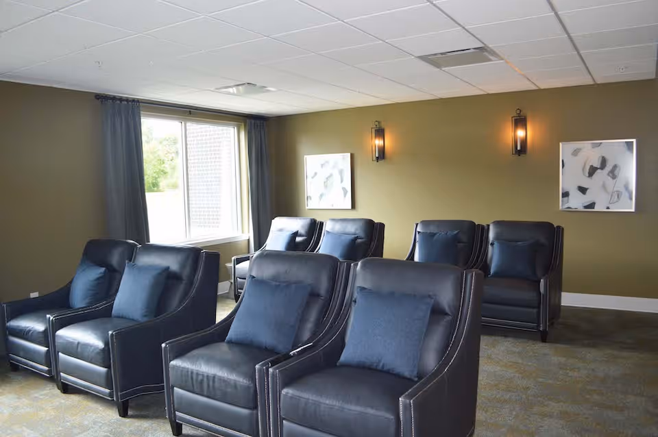A cozy sitting area with six black leather armchairs, each with a dark blue cushion, arranged in two rows facing forward. The room has olive green walls, two abstract paintings, two wall-mounted light fixtures, a window with dark curtains, and a carpeted floor.