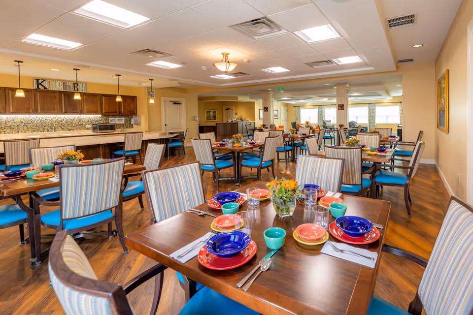 A bright and spacious dining room with multiple wooden tables set with colorful plates, bowls, cups, and silverware. Each table has a small vase with orange flowers. The chairs have striped upholstery with blue cushions. In the background, there is a kitchen area with wooden cabinets, a mosaic tile backsplash, and a sign that reads 'KITCHEN'. The room has warm lighting and wooden flooring.