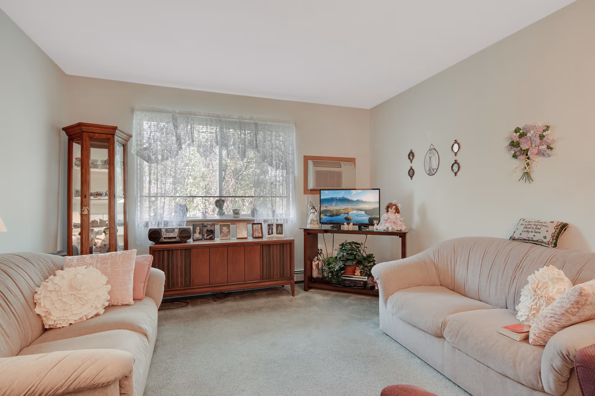 A cozy living room with two beige sofas adorned with decorative pillows, a wooden cabinet with framed photos and a stereo, a corner display cabinet with collectibles, a small table holding a TV and plants, and lace curtains covering a large window.
