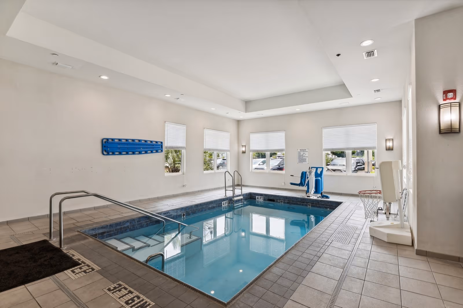 Indoor swimming pool area with tiled floor and walls, featuring a pool lift for accessibility, handrails, and steps leading into the pool. The room has several windows with blinds, allowing natural light to enter. There is a basketball hoop mounted on the wall near the pool.