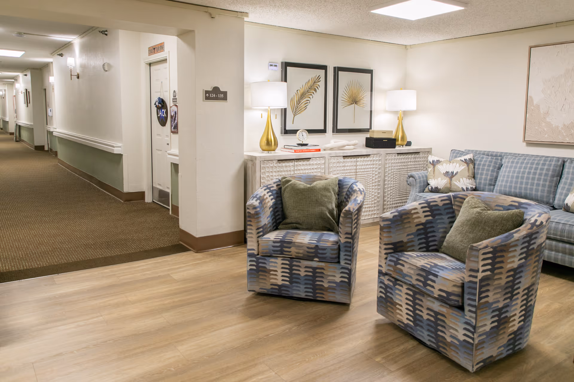 A cozy seating area in a senior living facility hallway with two patterned armchairs and a blue plaid sofa. Behind the chairs is a white textured cabinet with two gold lamps, decorative books, a clock, and framed botanical artwork on the wall. The hallway extends to the left with doors and wall-mounted lights.