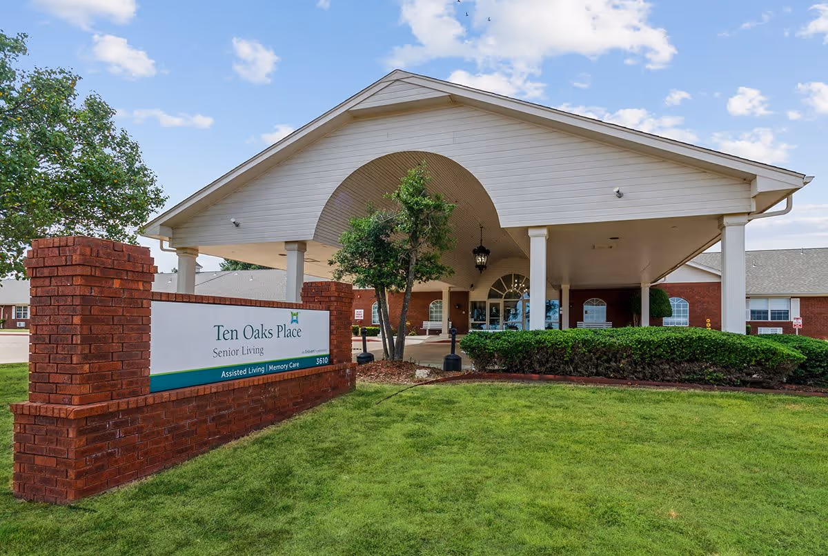 Exterior view of Ten Oaks Place Senior Living facility showing the entrance with a covered driveway, brick pillars, green bushes, and a sign indicating Assisted Living and Memory Care services.