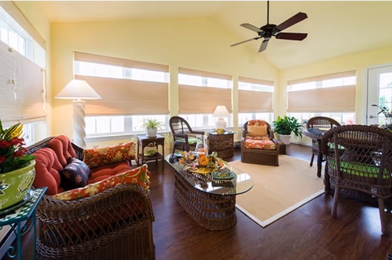 A bright and cozy living room with wicker furniture including a red cushioned sofa, armchairs with floral cushions, a glass-top coffee table, and a dining table with chairs. The room has large windows with beige blinds, a ceiling fan, hardwood floors, and several potted plants and lamps providing a warm atmosphere.