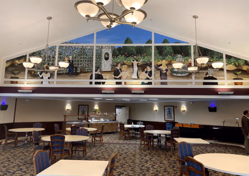 A spacious dining room with multiple round tables and chairs arranged on a patterned carpet. The room features high ceilings with hanging light fixtures and a large mural on the upper wall depicting people in formal attire dancing and socializing in a garden setting. The back wall has framed artwork, a buffet area, and a refrigerator. A person is standing near the right side of the room.