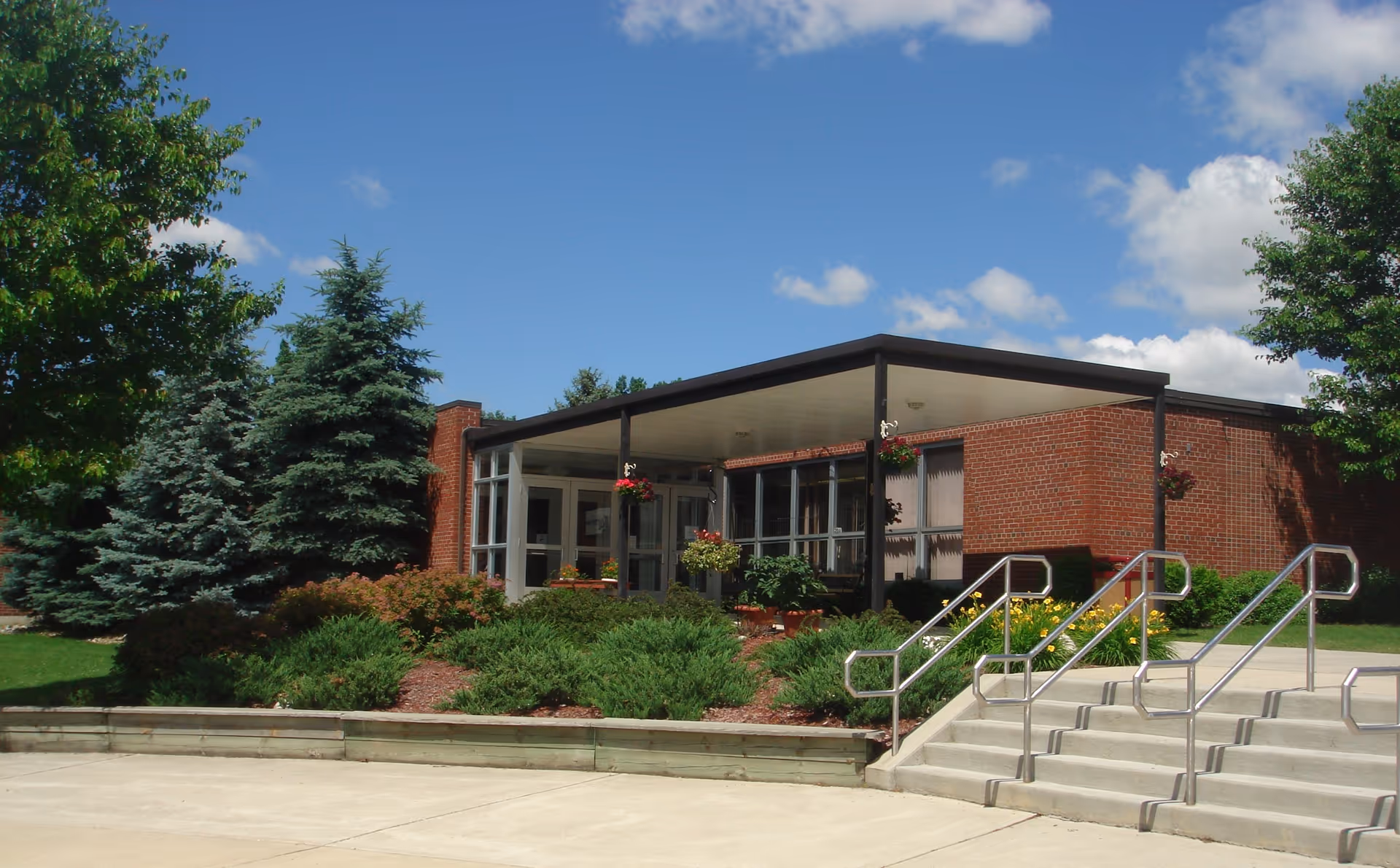 Low-rise brick building front with a covered entrance, steps and metal handrails, and landscaped shrubs and trees under a blue sky.