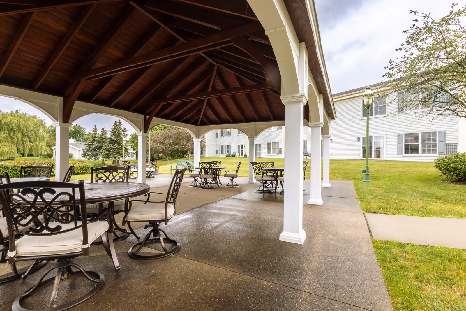 Outdoor covered patio area with multiple round tables and cushioned chairs under a wooden roof structure, surrounded by green grass, trees, and a white building in the background.