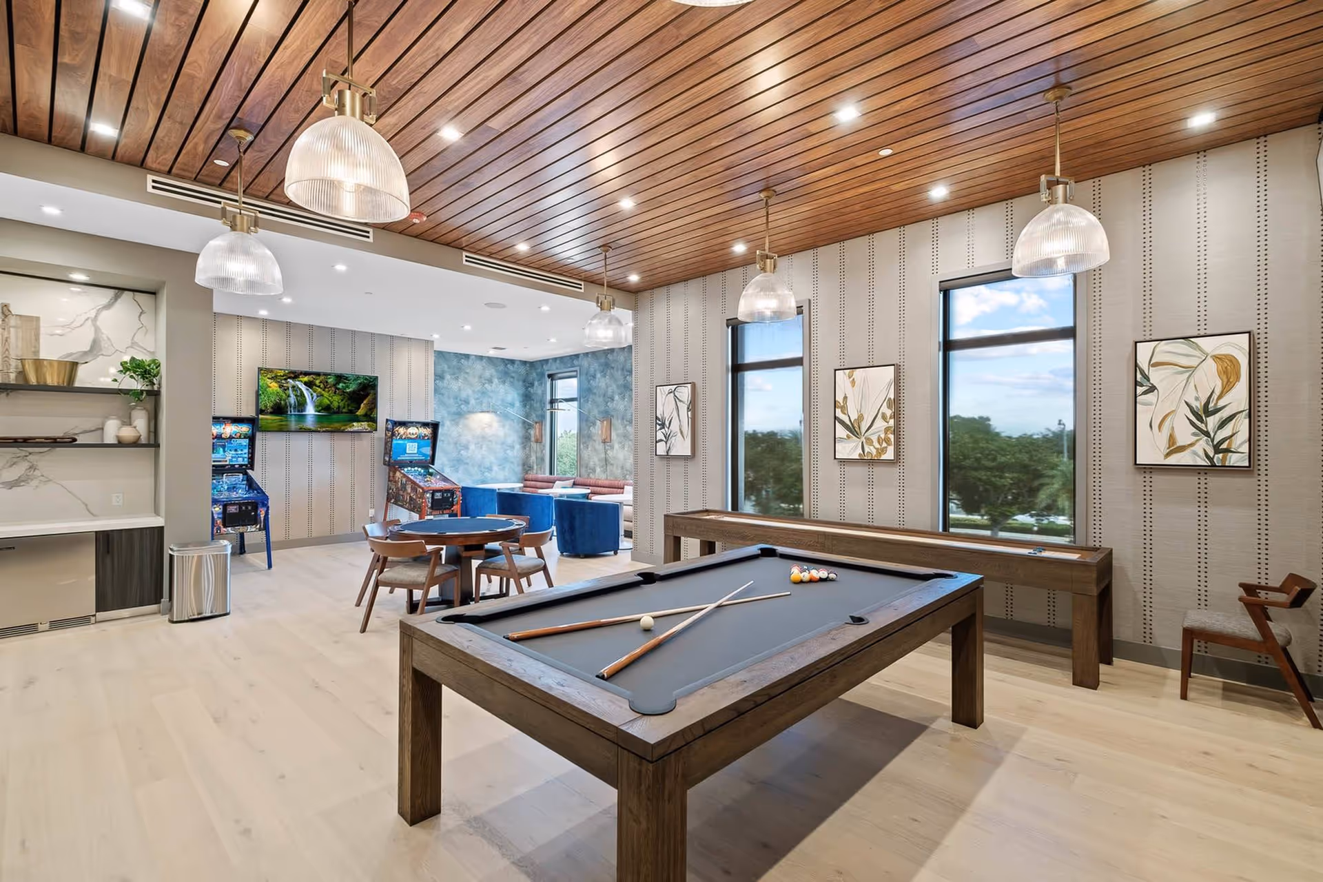 Bright recreation room with a pool table, shuffleboard, seating area, pinball machines, and large windows under a wood-paneled ceiling.
