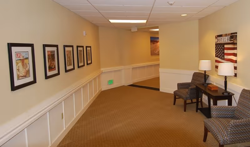 A hallway in a senior living facility with beige walls and carpeted floor. The left wall is decorated with framed vintage Life magazine covers, and the right side has two patterned armchairs, a small dark wooden table with two lamps, and a Route US 66 American flag wall art.