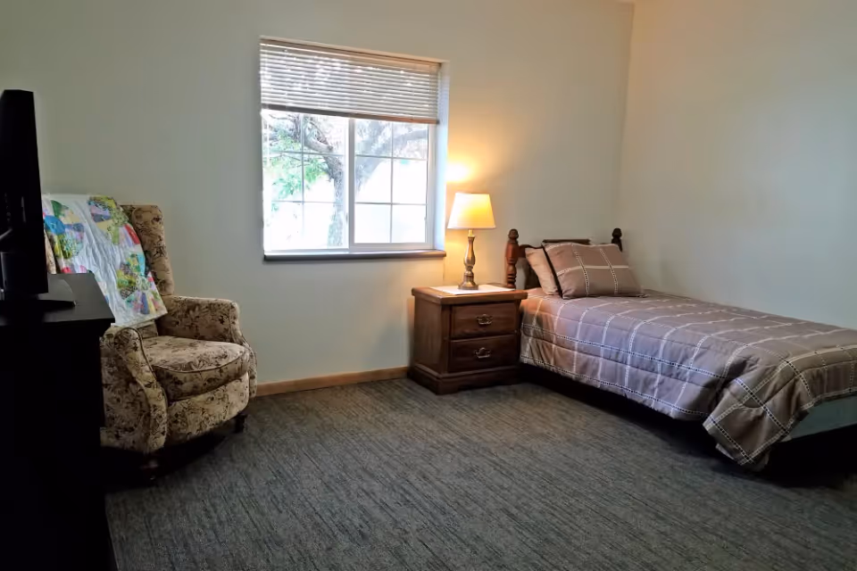 Single-occupancy bedroom with a twin bed, wooden nightstand and lamp, patterned armchair, and a window.