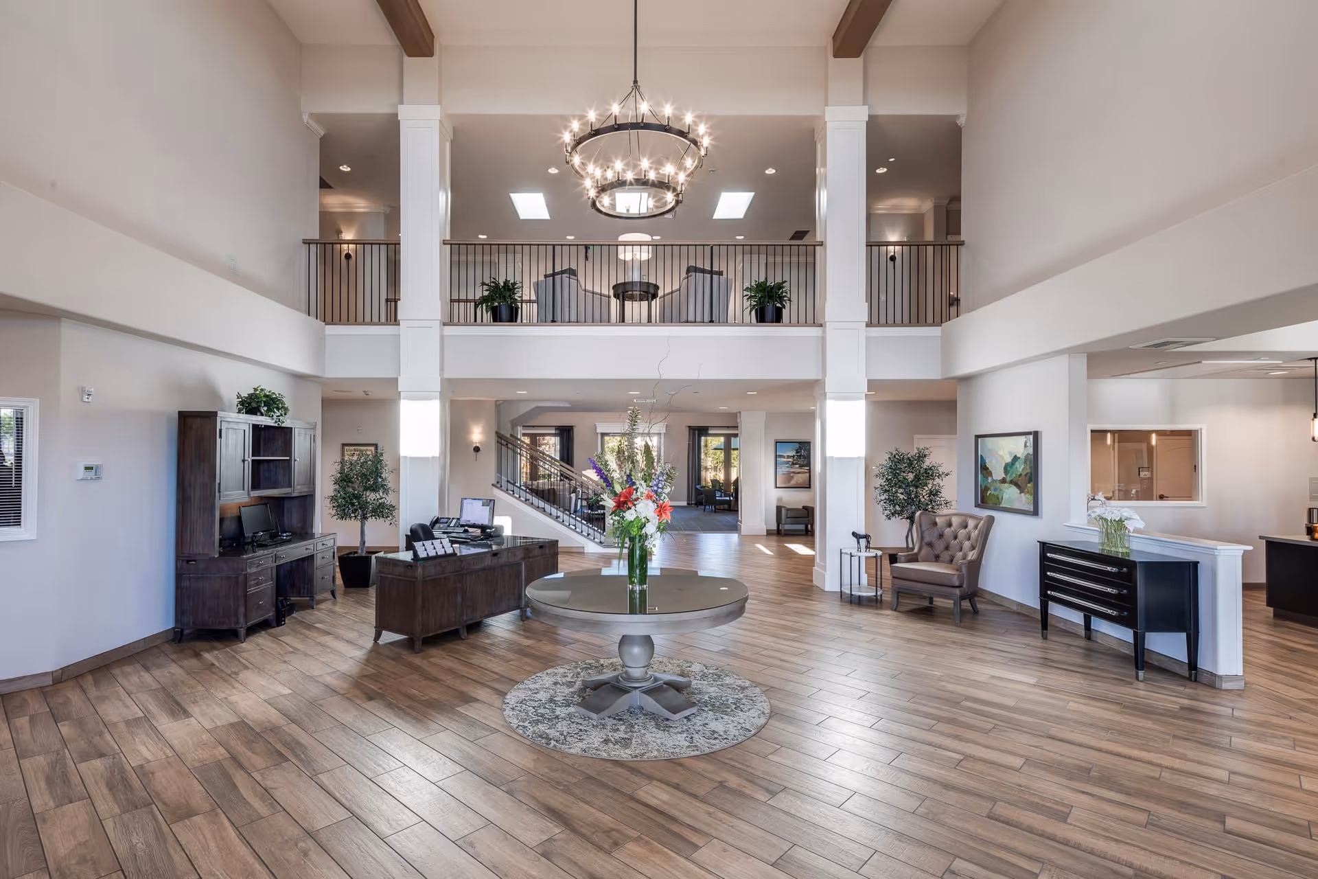 Spacious and well-lit senior living facility lobby with high ceilings, a large chandelier, wooden flooring, a round table with a flower arrangement in the center, a reception desk with computers, seating areas with chairs, potted plants, and artwork on the walls.