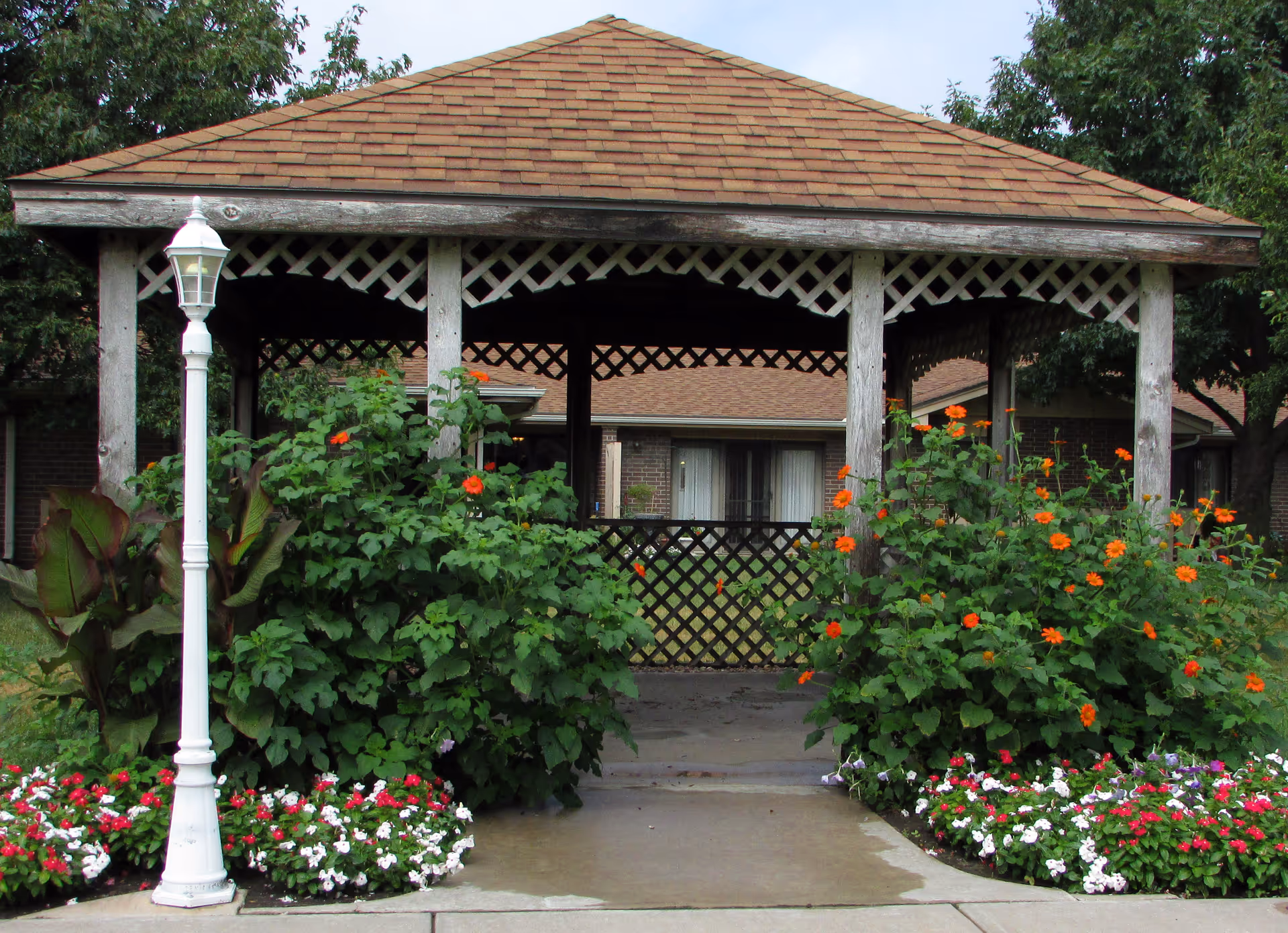 Wooden gazebo with lattice trim surrounded by flowering shrubs and a white lamp post in front of a brick building.