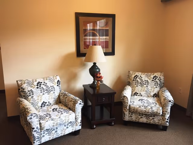 Two patterned armchairs with floral designs are positioned on either side of a dark wooden side table. The table holds a lamp with a beige shade and a small decorative item. A framed abstract artwork hangs on the beige wall behind the furniture.