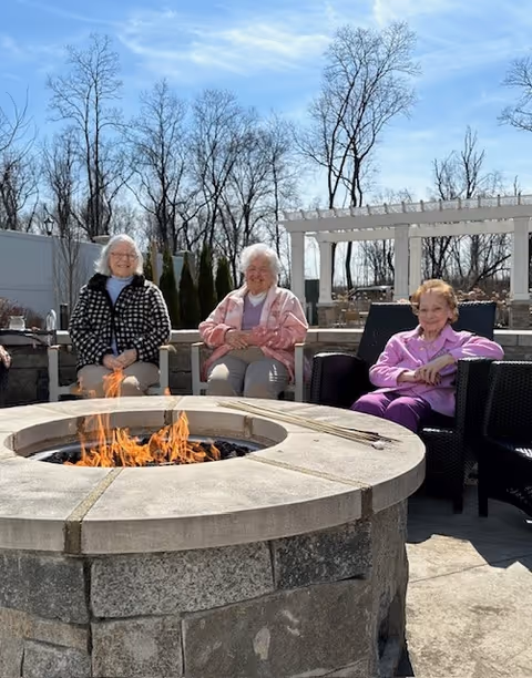 Three elderly women seated around a stone fire pit on an outdoor patio with leafless trees and a pergola in the background.